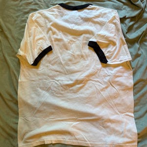 以下が含まれることがあります： 襟と袖に黒いトリムが付いた白いTシャツ。 Tシャツは緑色の表面に平らに置かれ、シンプルなデザインを示しています。 生地は柔らかく、わずかにしわが寄っているように見えます。