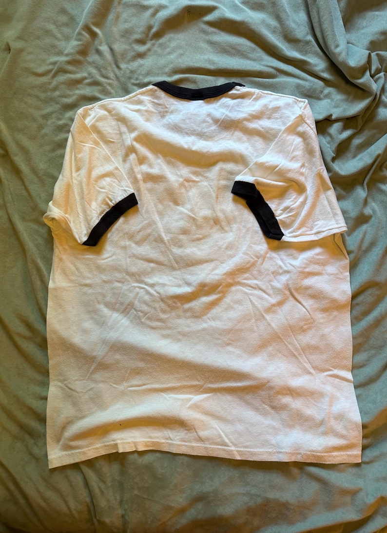 以下が含まれることがあります： 襟と袖に黒いトリムが付いた白いTシャツ。 Tシャツは緑色の表面に平らに置かれ、シンプルなデザインを示しています。 生地は柔らかく、わずかにしわが寄っているように見えます。