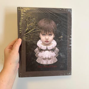 May include: A book wrapped in clear plastic featuring a portrait of a child with dark hair and a ruffled collar. The book's cover is dark with the text "The Distaff Side" in a serif font.
