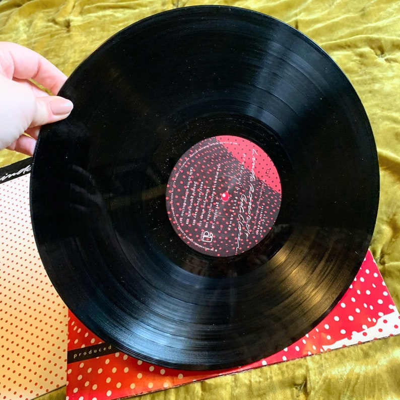 May include: Black vinyl record with a red and black label. The label has white polka dots and the text "Love & Rockets" in white. The record is on top of a red and white polka dot background.
