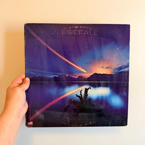 May include: Album cover for the band Firefall. The album art features a vibrant sunset over a lake with mountains in the background. A shooting star streaks across the sky. The album cover is held by a person.