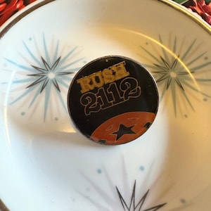May include: A round black and gold pin with the text "RUSH 2112" and a star design. The pin is set on a white bowl with a gold rim and a starburst pattern.