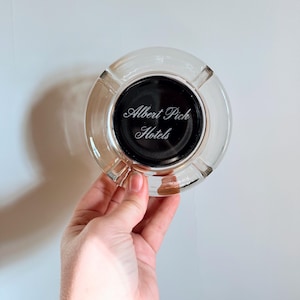 May include: Clear glass ashtray with a black center. The text "Albert Pick Hotels" is printed in white script on the black center. The ashtray has a circular shape with a wide rim.
