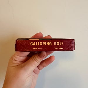 May include: A rectangular, burgundy-colored box with the words "GALLOPING GOLF" in gold lettering. The text "MADE IN U.S.A." and "PAT. PEND." are also visible. The box has a textured surface.