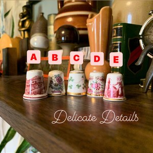 May include: Five porcelain thimbles with different designs. The thimbles are arranged in a row on a wooden surface. The thimbles are decorated with floral patterns, a building, and a woman in a dress. The thimbles are labeled A, B, C, D, and E. The text "Delicate Details" is written below the thimbles.