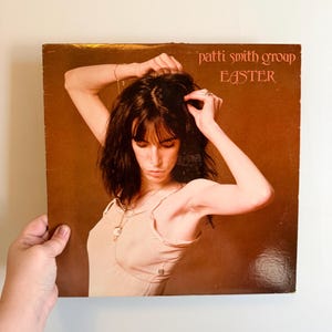 May include: A vintage vinyl record album cover featuring the text "patti smith group EASTER" in a reddish-brown font. The album cover has a sepia-toned image of a person with dark hair, wearing a light-colored top, with their arms raised.