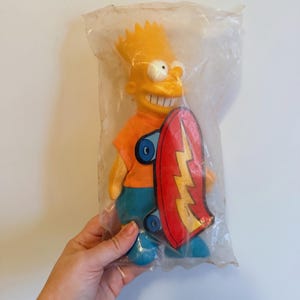 May include: A Bart Simpson plush toy in a plastic bag. The toy has yellow hair, an orange shirt, blue shorts, and blue shoes. He is holding a red and yellow skateboard with a lightning bolt design. The toy is a vintage collectible.