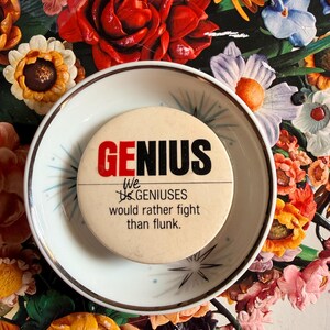 May include: A round, off-white button with the word "GENIUS" in red and black text. Below the word is the phrase "We GENIUSES would rather fight than flunk." The button rests on a small, white plate with a floral background.