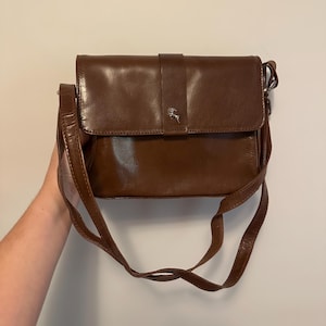 May include: A brown leather crossbody bag with a flap closure and a long, adjustable strap. The bag features a small, silver-colored emblem on the front. The bag is held by a person.