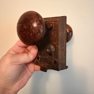 May include: An antique door lock with two brown, marbled doorknobs. The metal plate is rectangular and shows signs of rust. The doorknobs have a rounded, egg-like shape. The lock is held in a hand.