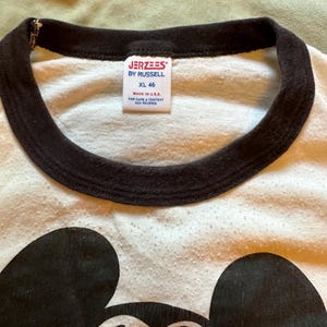 以下が含まれることがあります： 黒いミッキーマウスのグラフィックが入った白いTシャツ。シャツは茶色の襟で、タグには「JERZEES BY RUSSELL XL 46 MADE IN USA」と書かれています。