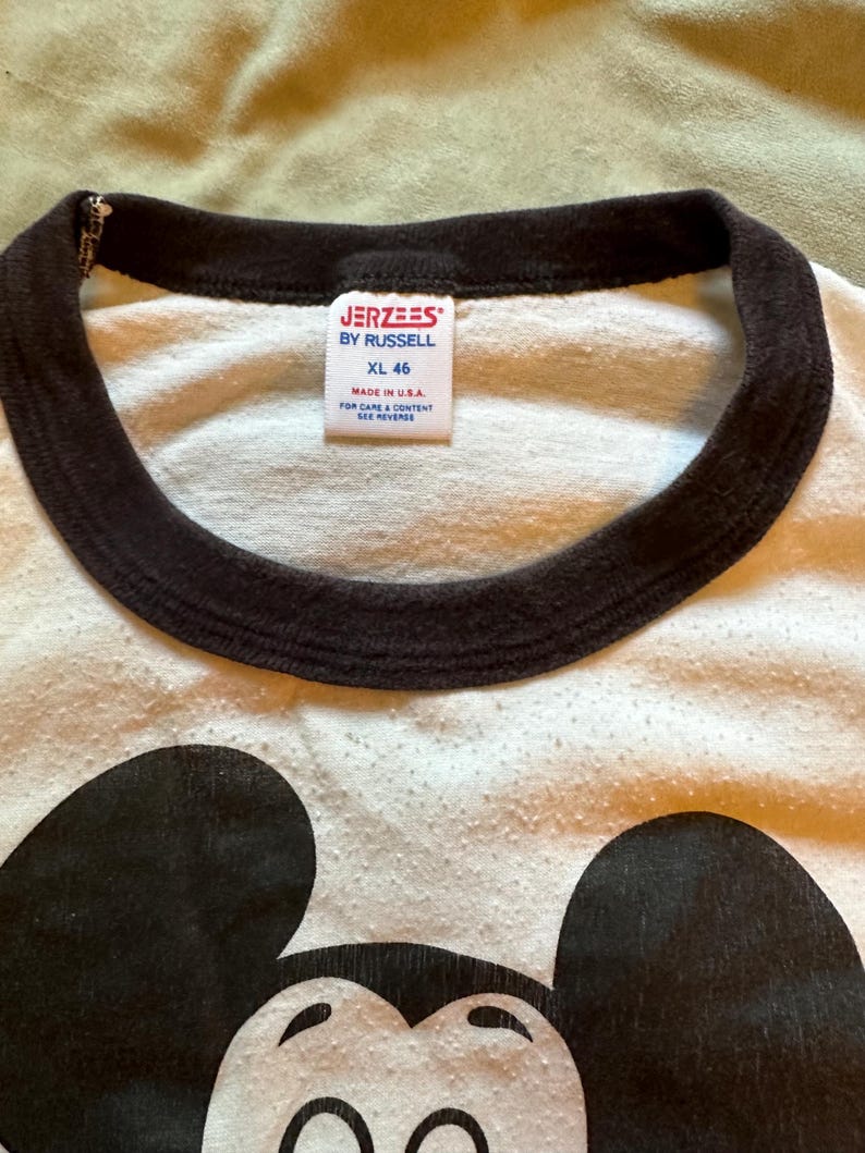 以下が含まれることがあります： 黒いミッキーマウスのグラフィックが入った白いTシャツ。シャツは茶色の襟で、タグには「JERZEES BY RUSSELL XL 46 MADE IN USA」と書かれています。