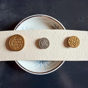 May include: Three vintage metal buttons with embossed text and designs. The buttons are in gold and silver tones, with one reading "Grand Rapids Railway Co." The buttons are arranged on a cream-colored strip, set against a dark background.