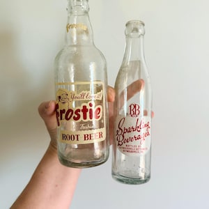 May include: Two vintage glass soda bottles. One bottle has a label that reads "Frostie Root Beer" with the text "You'll love it!". The other bottle has a label that reads "RB Sparkling Beverages".