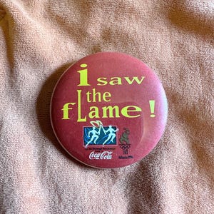 May include: A round, red button with the text "i saw the flame!" in yellow. The button also features the Coca-Cola logo and the Atlanta 1996 Olympic Games logo.