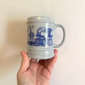 May include: A gray ceramic beer stein with a handle, featuring blue illustrations of landmarks and the text "Flamingo Hilton" and "Golden Nugget". The base is decorated with star-shaped designs.