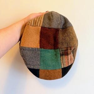May include: A patchwork newsboy cap featuring a variety of textured fabrics in shades of brown, gray, green, and burgundy. The cap is held up against a white background, showcasing its multi-colored design and classic style.