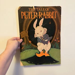 May include: Vintage copy of "The Tale of Peter Rabbit" book. The cover features an illustration of Peter Rabbit in a blue jacket and orange shoes. The title is in large orange letters. The book is held against a white wall.