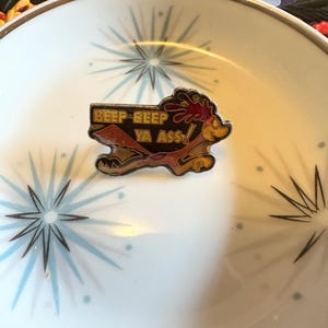 May include: A colorful enamel pin featuring the Road Runner cartoon character with the phrase "Beep Beep Ya Ass" in a rectangular banner. The pin is set against a white dish with a starburst design.