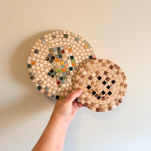 May include: Two mosaic coasters, one large and one small, held against a neutral wall. The coasters are circular, with a beige background and a colorful mosaic tile design. The larger coaster has a diameter of approximately 7 inches.