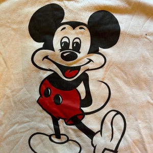 以下が含まれることがあります： ミッキーマウスの大きなグラフィックが特徴の白いTシャツ。漫画のキャラクターは黒、赤、白で描かれており、笑顔で腕を伸ばしています。シャツには黒い襟が付いています。