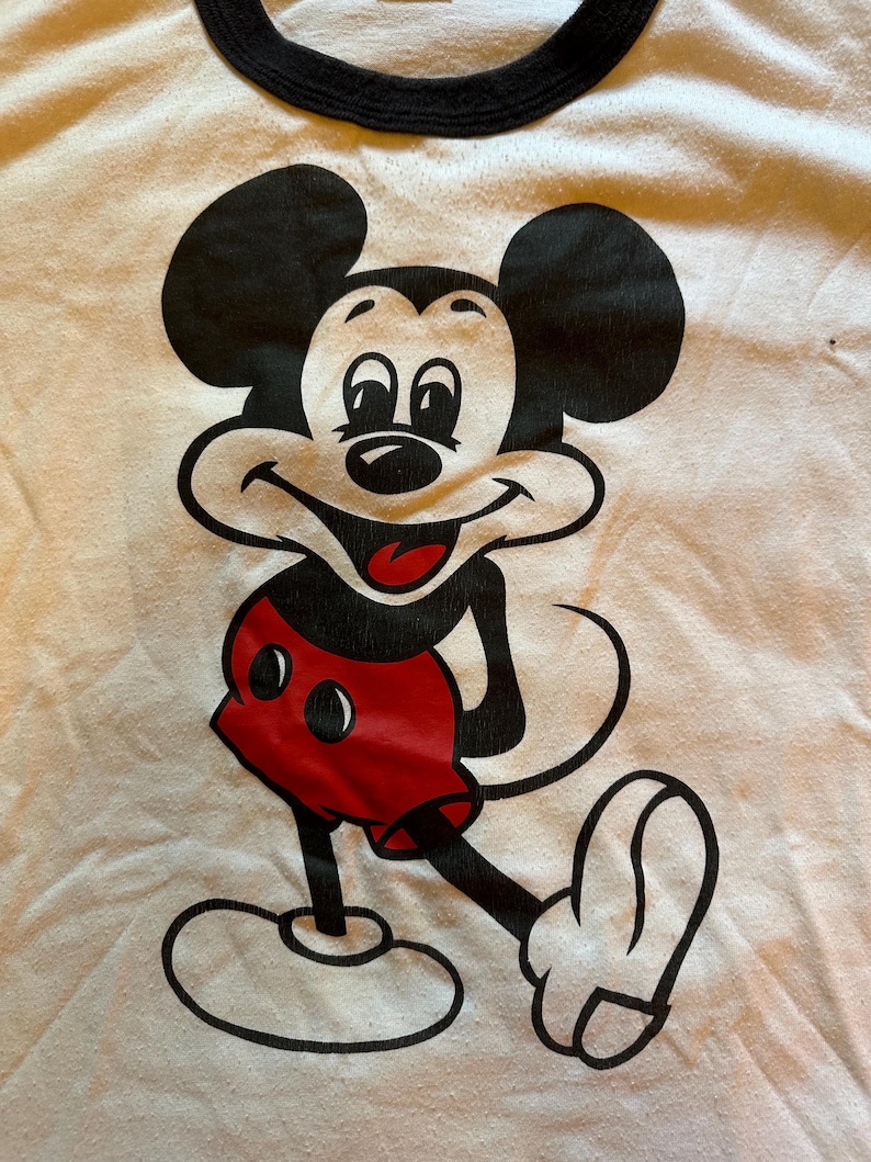 以下が含まれることがあります： ミッキーマウスの大きなグラフィックが特徴の白いTシャツ。漫画のキャラクターは黒、赤、白で描かれており、笑顔で腕を伸ばしています。シャツには黒い襟が付いています。