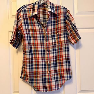 May include: A short-sleeved plaid shirt in a classic button-down style. The shirt features a vibrant pattern of red, blue, yellow, and white checks. It has a chest pocket and is hanging on a hanger against a white door.