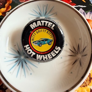May include: A vintage Hot Wheels button featuring a blue car graphic with the text "Nitty Gritty Kitty" and "Mattel Hot Wheels." The button is set on a white and gold rimmed dish with blue starburst designs.