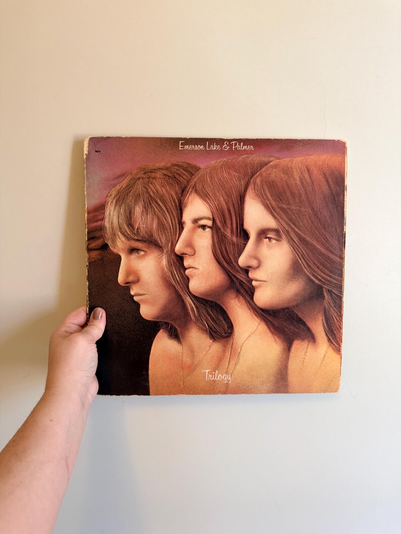 May include: Vintage vinyl record album cover for Emerson, Lake & Palmer's "Trilogy." The cover art features three side profiles of people with long hair, in shades of brown and gold. The album title and band name are printed on the cover.