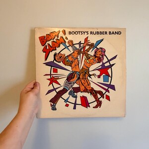 May include: Vintage vinyl record album cover for Bootsy's Rubber Band. The album art features two cartoon figures in action, surrounded by geometric shapes in red, blue, and purple. The album title is displayed at the top.