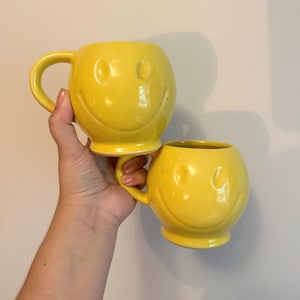 May include: Two yellow ceramic mugs with smiling faces. Each mug has a rounded shape, a handle, and a pedestal base. The mugs are held in a person's hand against a plain white background.