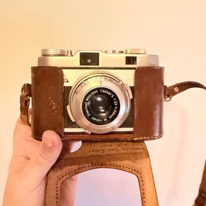 May include: Vintage silver and black camera with a brown leather case. The camera lens reads "Cassar S 1:28 f=50mm" and "Steinheil München". The case has a strap and is open, revealing the camera's front.