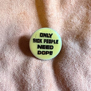 May include: A round, yellow button with the text "ONLY SICK PEOPLE NEED DOPE" in black, bold letters. The button is centered on a peach-colored, textured surface.