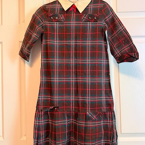 May include: A long-sleeved dress featuring a dark green, red, and white plaid pattern. The dress has a cream-colored collar with a small red bow at the center, two decorative tabs with buttons on the chest, and a pleated skirt section at the hem.