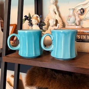 May include: Two turquoise ceramic mugs with ribbed sides and large handles sit on a wooden shelf. The mugs are part of a vintage collection, displayed alongside decorative figurines and books. The mugs are a bright, solid color.