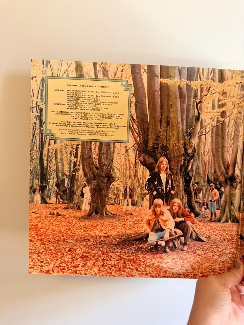 May include: Vintage vinyl record album cover for Emerson, Lake & Palmer's "Trilogy." The cover art features a forest scene with the band members. The album title and track list are visible on the cover. The overall color palette is warm, with shades of brown and orange.