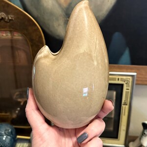 May include: A light brown ceramic vase shaped like a teardrop with a small opening at the top. The vase has a smooth, glossy finish.