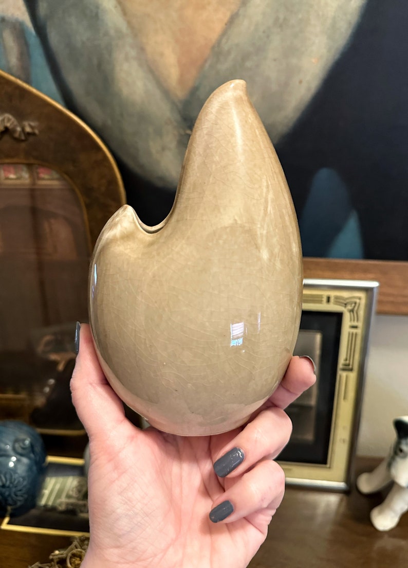 May include: A light brown ceramic vase shaped like a teardrop with a small opening at the top. The vase has a smooth, glossy finish.