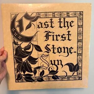 May include: A square vinyl record jacket with a light tan background featuring black ink illustrations. The text reads Cast the First Stone. Syn in a gothic calligraphy font, surrounded by a decorative border of leaves and geometric patterns.