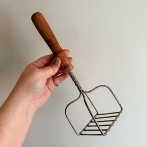 May include: A vintage potato masher with a wooden handle and a metal mashing head. The handle is a warm brown color, and the metal head has a square shape with parallel wires. The masher is designed for kitchen use.