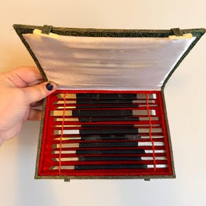 May include: A set of carving tools in a decorative box. The box is green with a patterned design and a red velvet interior. The tools have black handles and silver blades. The box is open, revealing the tools neatly arranged.