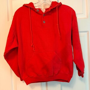 May include: A bright red long-sleeve hooded sweatshirt featuring a three-button placket at the neckline, a front kangaroo pocket, and matching red drawstrings. The garment includes a small white label at the collar with the text GAP printed in black.
