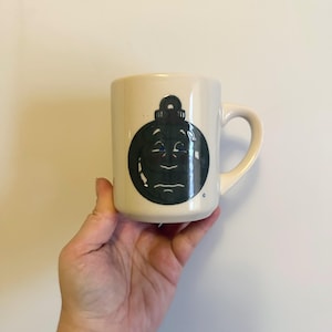 May include: Cream-colored ceramic mug featuring a black ornament design with a smiling face. The mug has a curved handle and a cylindrical shape. The ornament design is centered on the mug's front.