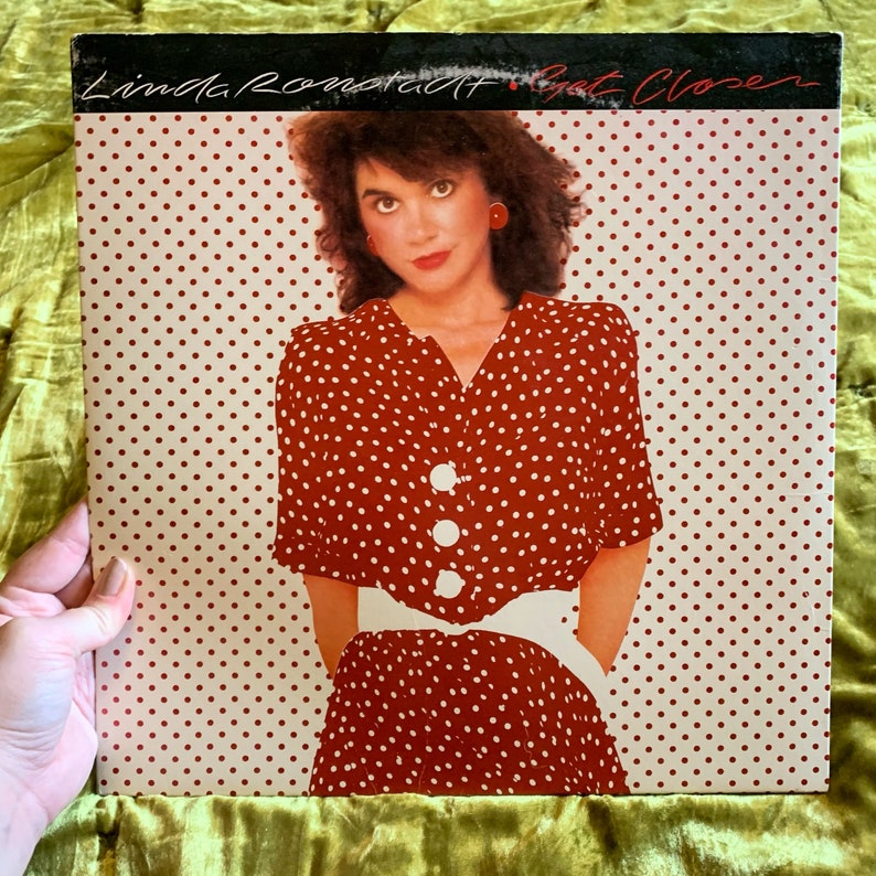 May include: A red and white polka dot dress with a white belt and white buttons. The album cover features the text "Linda Ronstadt - Get Closer".