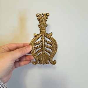 May include: A decorative brass wall hanging with an ornate, symmetrical design. The piece features a central floral motif with stylized leaves and a loop at the top for hanging. The brass has a warm, aged patina.