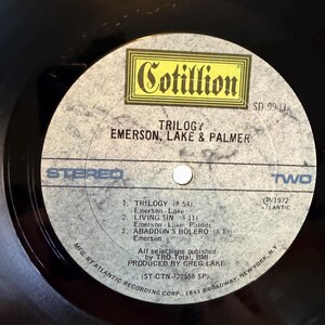 May include: A black vinyl record with a gray and yellow label. The label features the text "Cotillion" and "Trilogy Emerson, Lake & Palmer." The tracklist includes "Trilogy," "Living Sin," and "Abaddon's Bolero." The record is a stereo recording.