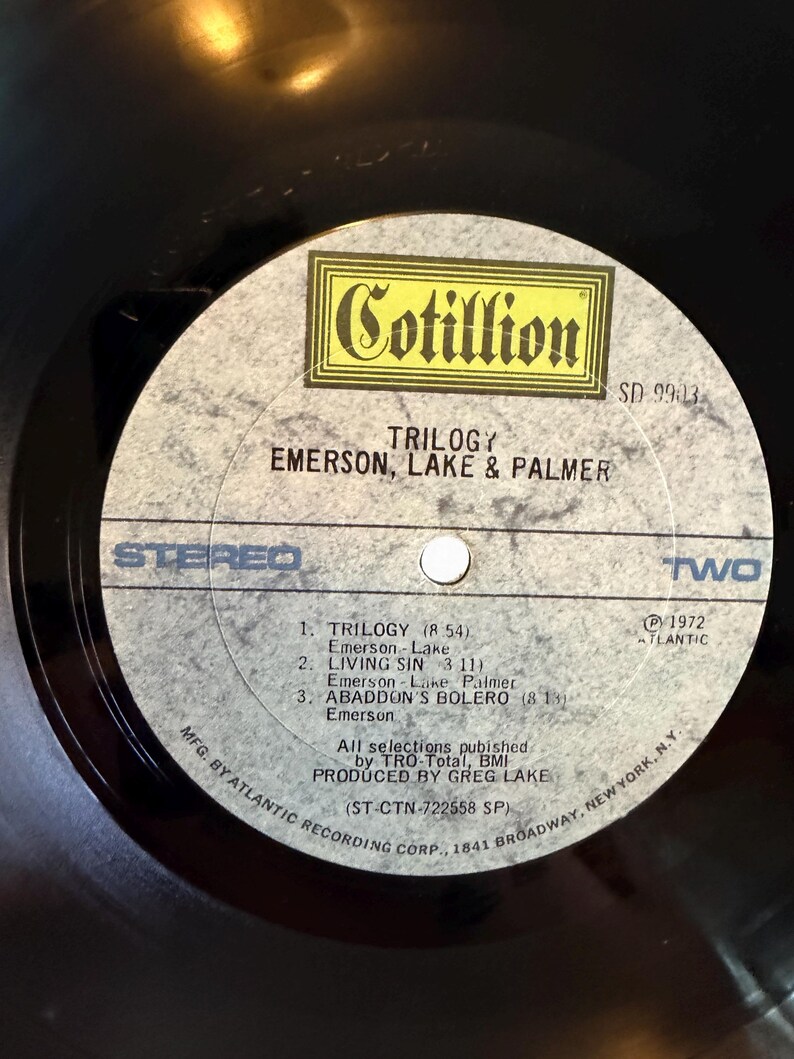 May include: A black vinyl record with a gray and yellow label. The label features the text "Cotillion" and "Trilogy Emerson, Lake & Palmer." The tracklist includes "Trilogy," "Living Sin," and "Abaddon's Bolero." The record is a stereo recording.