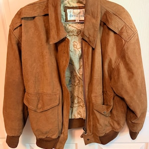 May include: A brown suede bomber jacket with a collar, long sleeves, and ribbed cuffs and hem. The jacket features two front flap pockets with snap closures and a front zipper. The interior lining displays a printed map design and a Donna Pell label.
