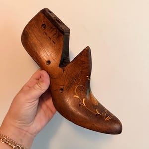May include: A dark brown wooden shoe last featuring a pointed toe and a high heel section. The surface is decorated with hand-painted gold floral scrollwork and the word WELCOME written in gold lettering near the toe area of the wooden form.