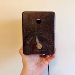 May include: A vintage brown metronome with a dial and a pendulum. The metronome is held in a hand against a white background. The word "Metronoma" is printed on the front.